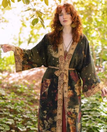 Person in dark floral robe with fox and moon design standing in sunlit garden.