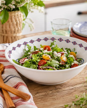 White patterned serving bowl filled with salad on a rustic wooden table with wooden utensils and greenery.