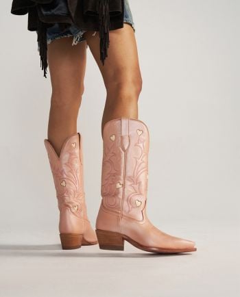 Woman wearing tall pink cowboy boots with heart cutouts, shown from mid-thigh down against a plain background.