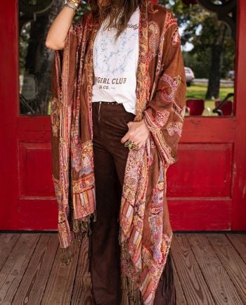 Woman wearing a patchwork paisley kimono over a graphic tee and brown fringe pants in front of red doors.