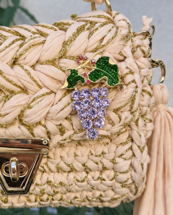 Cream woven handbag with gold accents and a jeweled grape brooch featuring purple stones and green leaves.