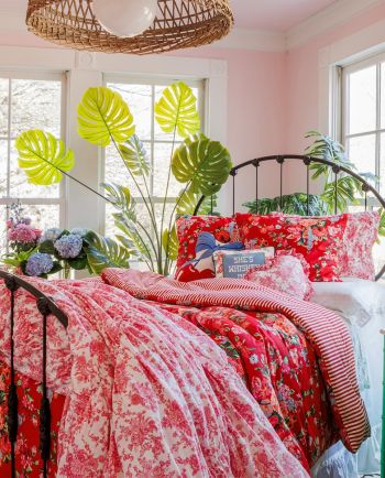 Bright bedroom with red floral bedding, striped throw, black metal bed, and large green plants by windows.