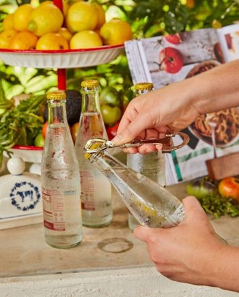 Hands opening a glass soda bottle with a metal opener beside other bottles, lemons, and a cookbook on a counter.