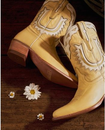 Cream western boots with stitched detailing arranged on dark wood beside small white daisies.