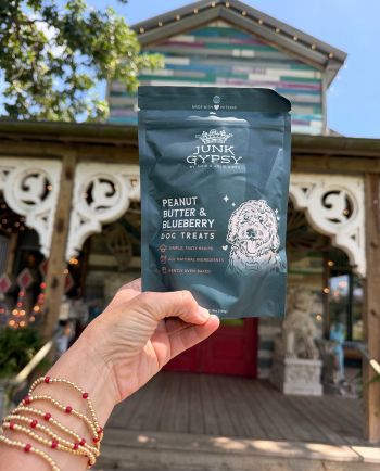 Hand holding a bag of Junk Gypsy peanut butter and blueberry dog treats in front of a colorful Texas storefront.