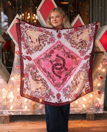 Woman holding pink floral scarf with horses and horseshoe design in front of lit vintage sign.