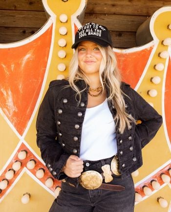 Smiling woman in black denim jacket and graphic tee with leopard-print pants standing in a doorway.