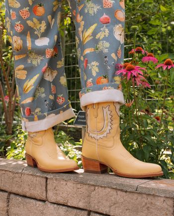 Cream western boots with stitched detailing arranged on dark wood beside small white daisies.