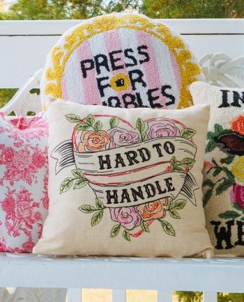 Throw pillows on a bench, featuring floral designs and text including “Hard to Handle” and “Press for Bubbles.”