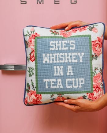 Hands holding a floral needlepoint pillow that reads “She’s whiskey in a tea cup” against a pink background.