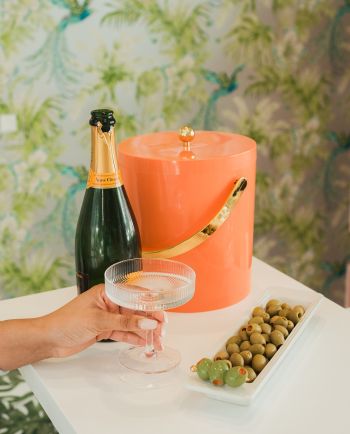 Hand holding a coupe glass beside champagne, an ice bucket, and a tray of green olives with a decorative olive pick.