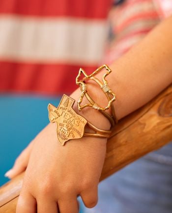 Close-up of gold-tone wire cuff bracelets featuring a Texas state outline and engraved Texas-shaped charm.