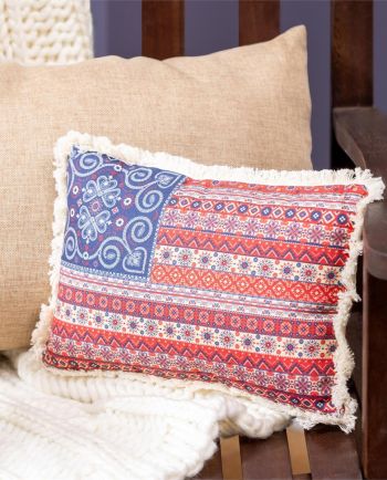 Rectangular red and navy patterned fringe pillow resting on a wooden bench with neutral pillows and a white throw blanket